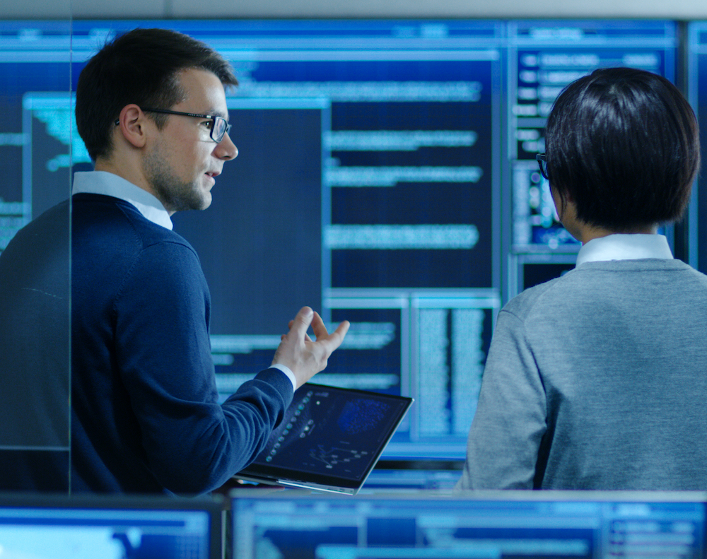 In the System Control Room IT Specialist and Project Engineer Have Discussion while Holding Laptop, they're surrounded by Multiple Monitors with Graphics. They Work in a Data Center on Data Mining, AI and Neural Networking. CITOC